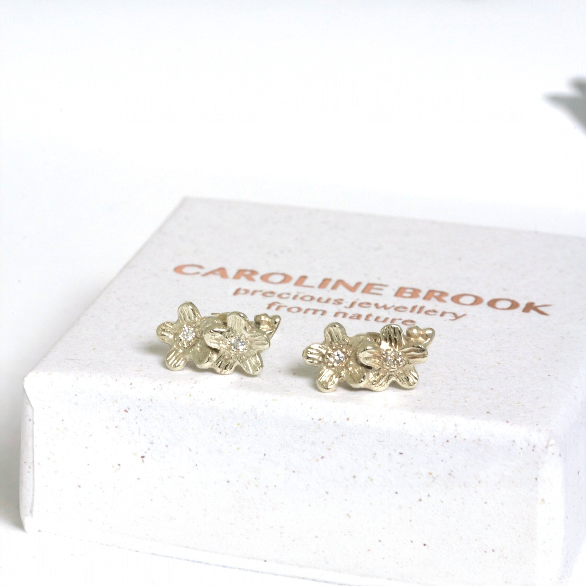 Gold floral earrings on a white box with 'Caroline Brook' branding.