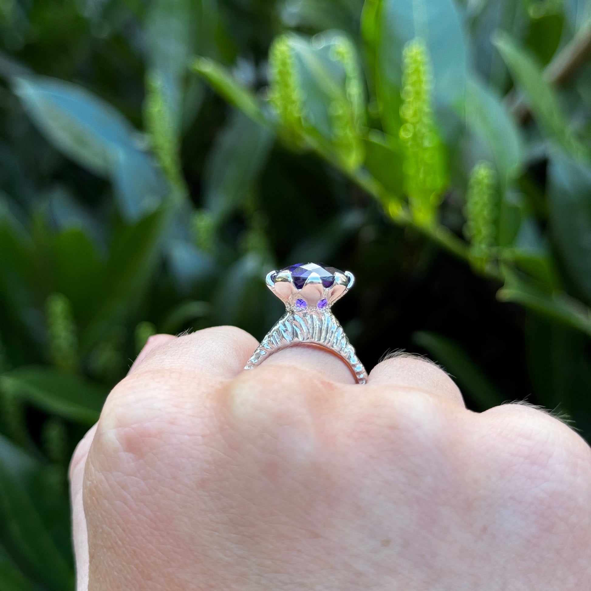 Hand-carved sterling silver ring with a high tulip petal-style bezel holding a dark purple oval chequerboard-cut amethyst, sculptural carved shank and elegant raised setting.