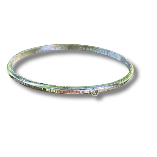A hammered sterling silver bangle bracelet with a small round peridot gemstone accent on the front, shown on a white background with a soft drop shadow.