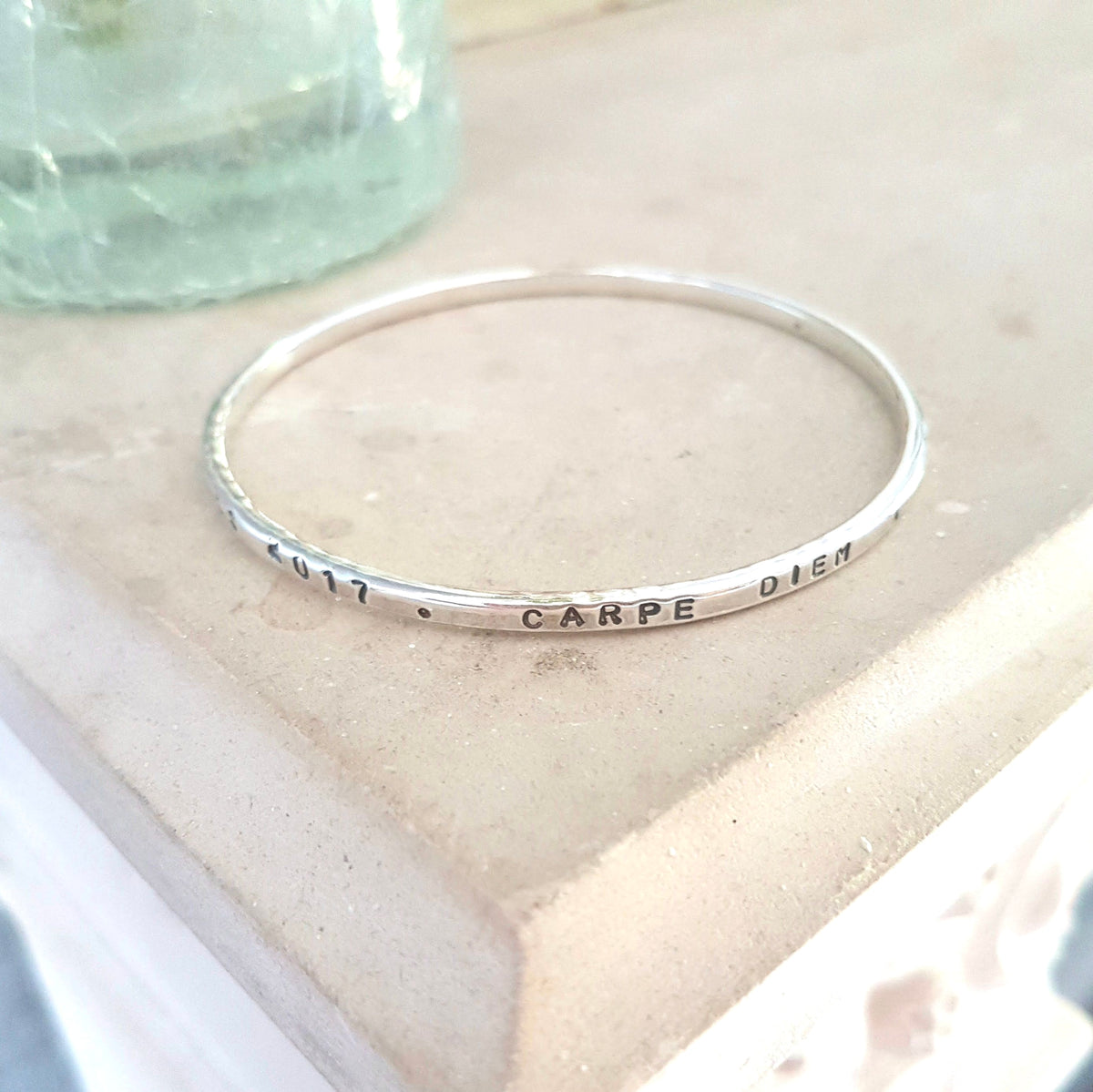 A slim silver bangle bracelet with the words 'CARPE DIEM' and date hand-stamped on the band, resting on a textured beige stone surface.