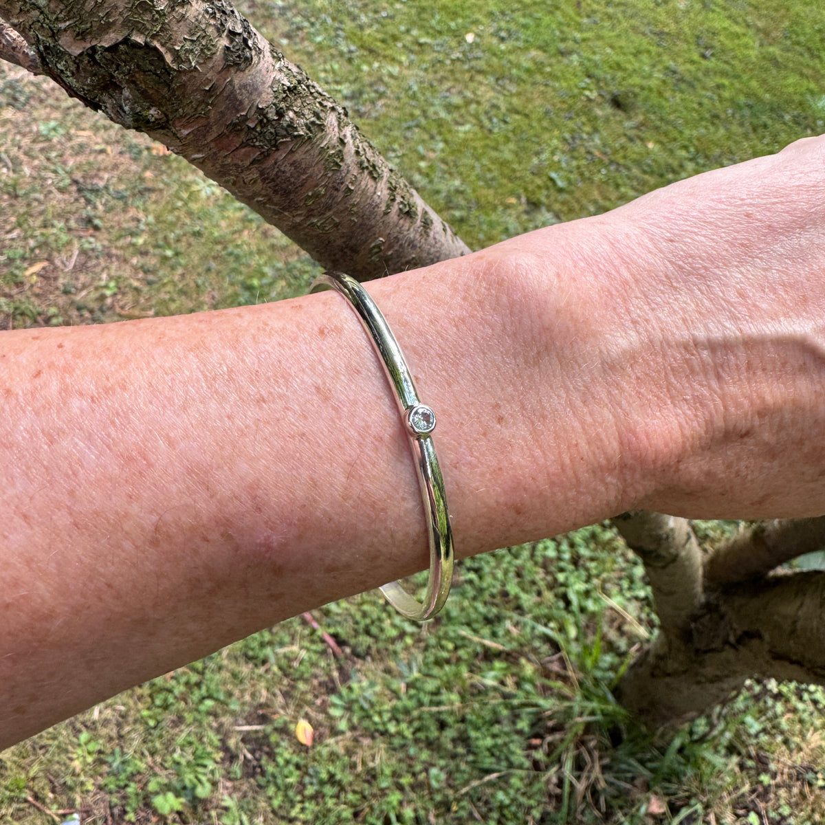 A polished silver bangle bracelet with a central clear stone, worn on a person's wrist outdoors against a background of grass and tree branches.