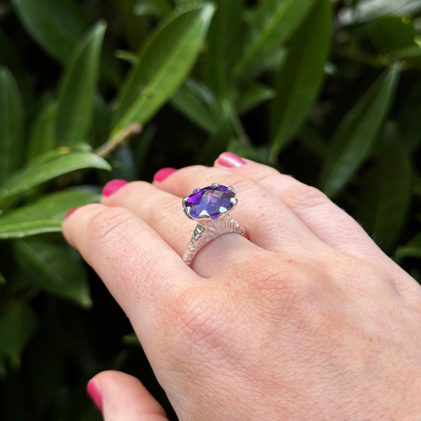 Hand-carved sterling silver ring with a high tulip petal-style bezel holding a dark purple oval chequerboard-cut amethyst, sculptural carved shank and elegant raised setting.