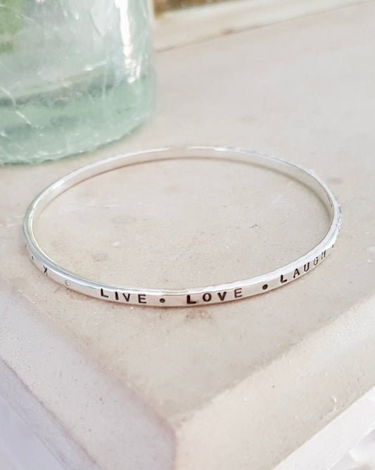 A thin silver bangle bracelet with the words 'LIVE LOVE LAUGH' stamped  on the band, resting on a light beige stone surface next to a textured glass jar.