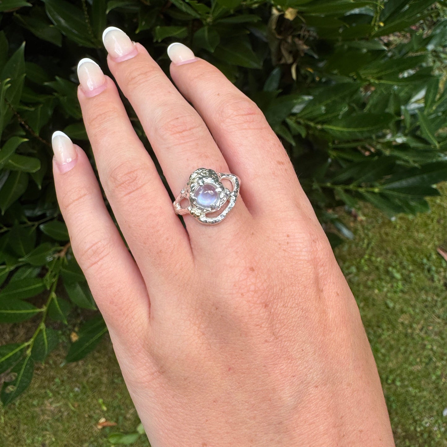 Close-up of a hand-carved silver ring set with a round blue moonstone, accented with 18 carat gold balls and gold inlay on an organic textured band.