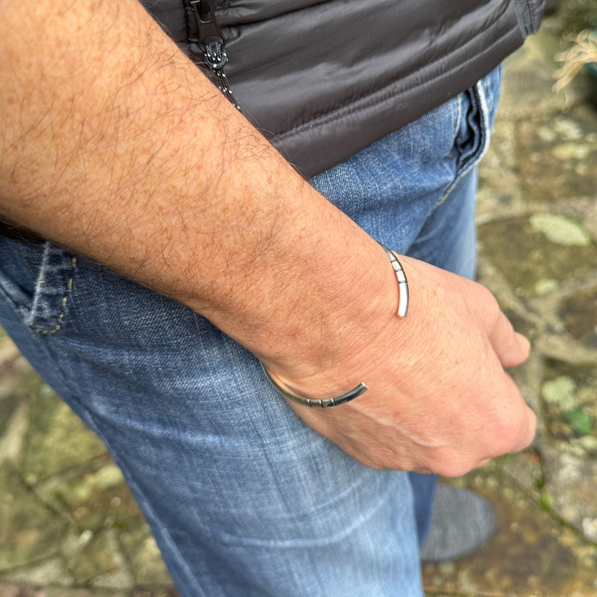 A close-up of a person's forearm and hand wearing a cuff silver bracelet outdoors, with blue jeans and a dark jacket visible in the background.