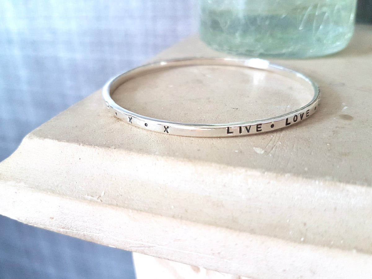 A slim silver bangle bracelet with engraved text and symbols, resting on a light-colored stone surface.