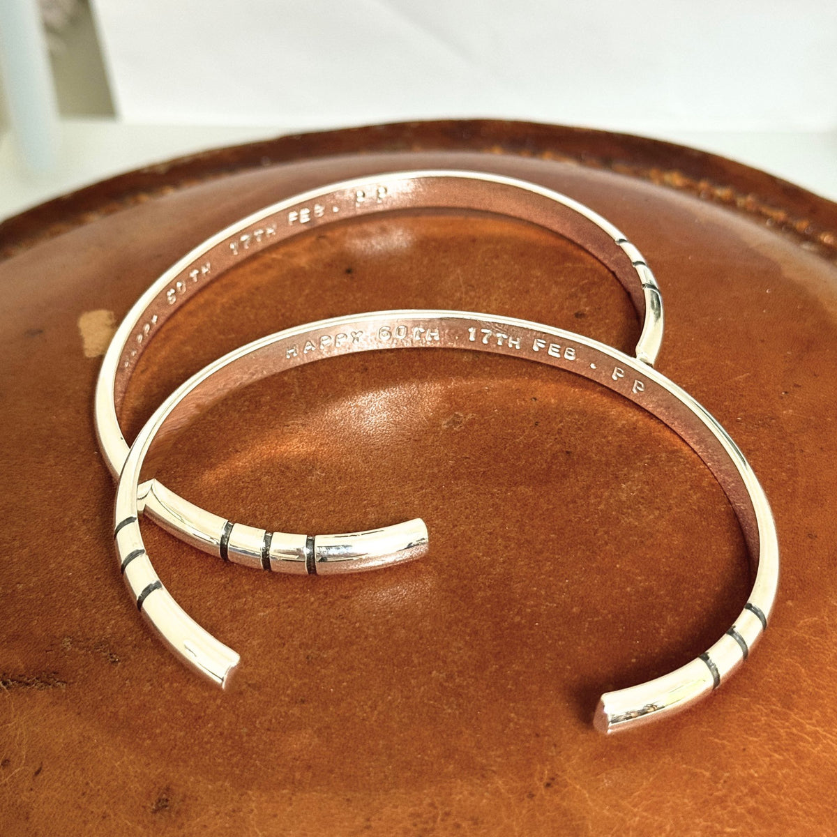 Two polished silver cuff bracelets with engraved text and black stripe details, displayed on a brown leather surface.