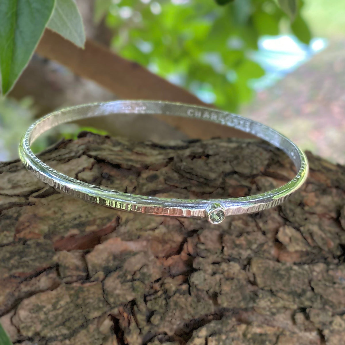 A textured sterling silver bangle bracelet with a small round bezel-set clear stone, resting on a piece of tree bark outdoors with green leaves blurred in the background. The inside of the bracelet is personalised  with the name 'CHARLOTTE'