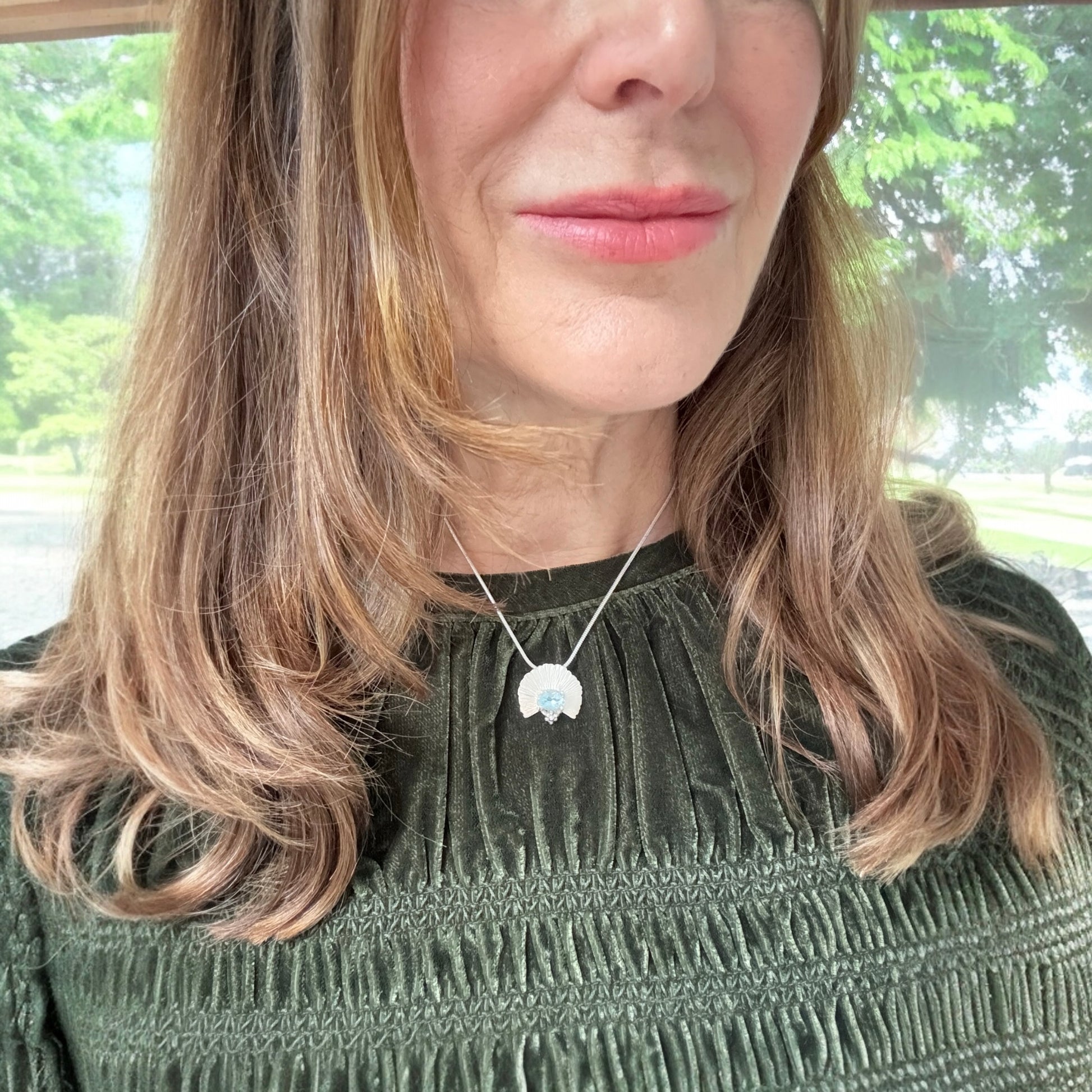 Woman wearing a necklace with a fan pendant in front of a window with greenery.