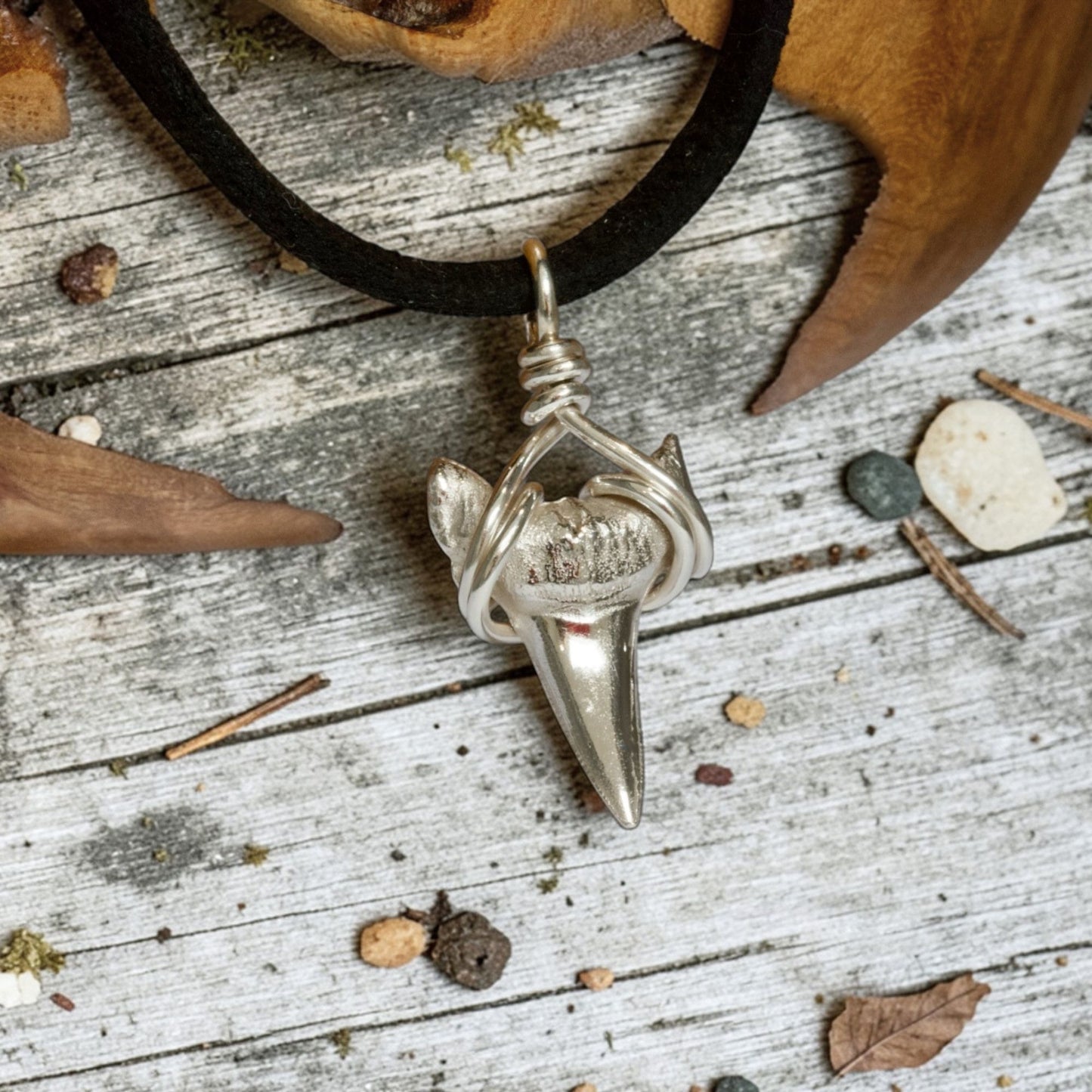 Solid sterling silver shark's tooth pendant on a faux suede thong necklace on a beach boardwalk with scattered pebbles and shells.