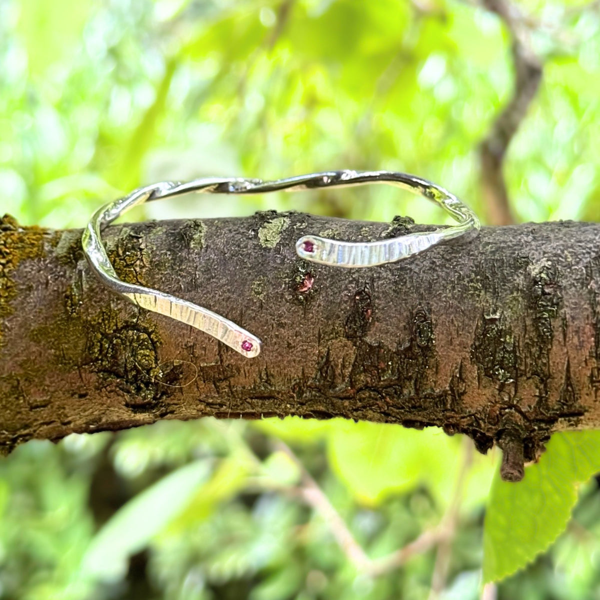 A silver open cuff bracelet with two textured, hammered bar-like ends and small red gemstone accents, resting on a mossy tree branch with a blurred green forest background.